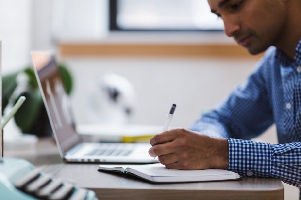 man at desk writing in notebook, with laptop open beside him