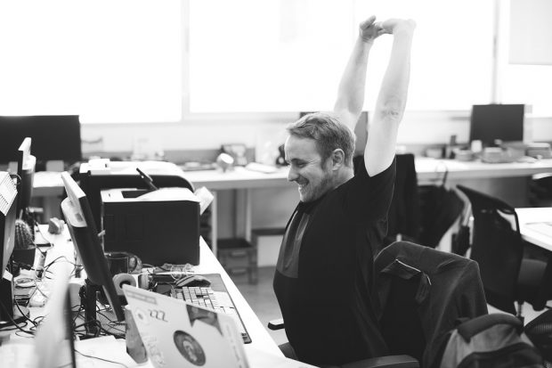 man in office, working on desktop PC, smiling, arms in the air, in expression of joy.